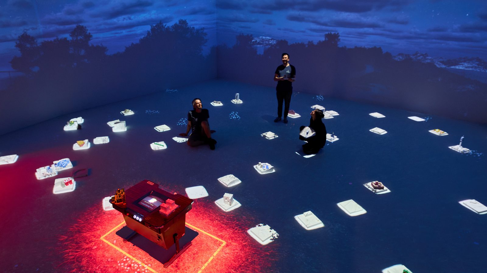 Three visitors kneel among illuminated miniature buildings spread across the lab floor, around a red-lit console.
